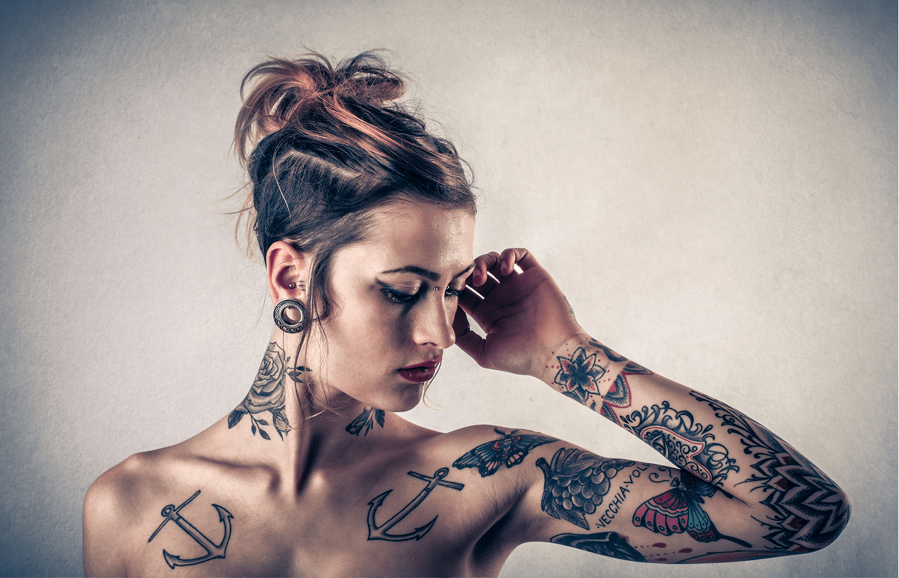 A tattooed woman poses with a contemplative expression, showcasing intricate body art on her arms and neck. The neutral background highlights her style.